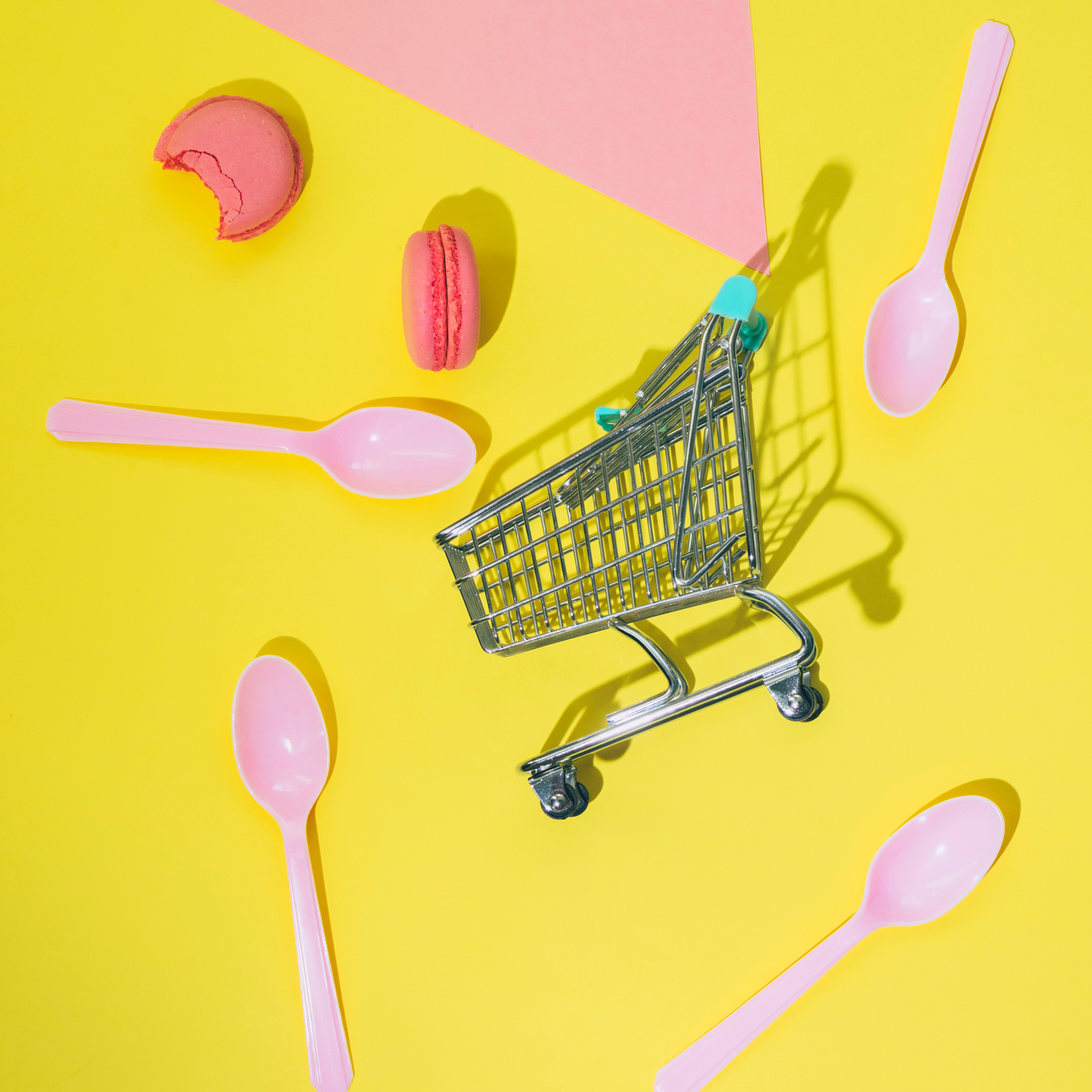 Pink almond cookies macarons or macaroons fall from shopping cart with plastic spoons on yellow and pink background.